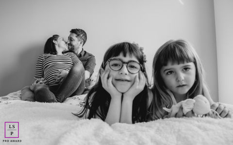 The black and white tones masterfully highlight a family in transition, layering the girls' curious foreground gazes against their parents' quiet, expectant embrace. It is a witty, heartwarming study of home.