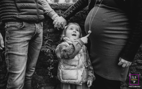 The child’s wide-eyed wonder provides a delightful centerpiece as she points toward the mystery of her mother’s pregnant belly. This black and white frame masterfully captures a beautiful, curious family threshold.