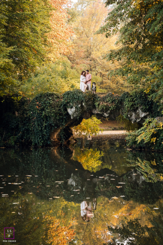 Surrounded by autumn color in Lyon, this expecting couple stands with their dog atop a vine covered bridge reflected perfectly below. The layered symmetry and peaceful landscape give the photograph romance, stillness, and storybook charm.