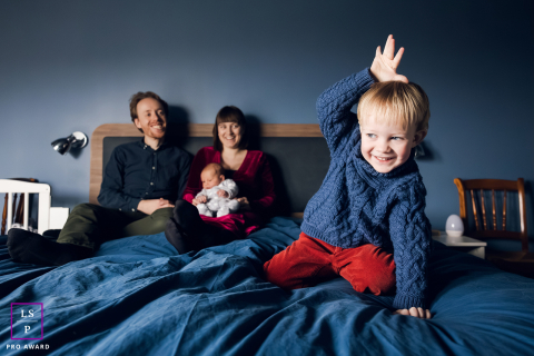 In this Lyon bedroom, the oldest son completely steals the scene with playful chaos while his parents and newborn laugh from the background. The layered composition and deep blue tones turn ordinary family life into something lively and honest.