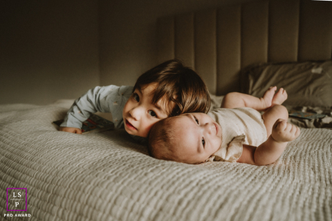 In this softly lit Toronto bedroom, a young brother presses close beside his baby sister with effortless tenderness. The warm shadows and low perspective make the photograph feel intimate, peaceful, and beautifully centered on quiet sibling connection.