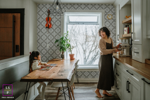 In this Toronto kitchen, a mother pauses mid task to share a warm glance with her daughter across the table. The patterned walls, soft window light, and everyday setting give the photograph comfort, rhythm, and quiet domestic beauty.