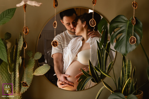 Expecting Couple Framed By Plants And Mirrored Calmly In Tender Châteauroux, France Maternity Portrait In Châteauroux, this expecting couple is framed by plants and reflected softly in the mirror, creating a photograph filled with calm anticipation. The layered composition and warm interior tones give the scene intimacy, tenderness, and natural elegance.