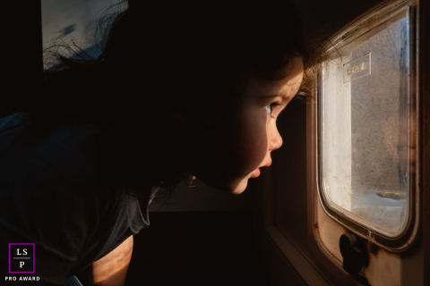 Young Girl Peers Through Pet Door With Focused Curiosity And Warm Light In Indre, France Portrait In Indre, a young girl peers through the small pet door with complete concentration, as though discovering another world beyond the glass. The warm side light and tight framing transform simple curiosity into something cinematic and deeply human.