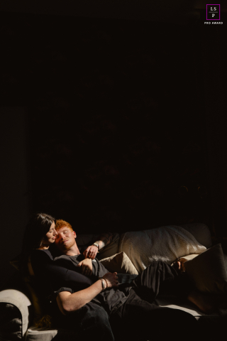 Couple Sinks Into Couch Bathed In Painterly Evening Light For Quiet Châteauroux, France Portrait In Châteauroux, this couple sinks quietly into the couch as late light cuts through the darkness with painterly precision. The deep shadows and narrow band of illumination make the photograph feel intimate, cinematic, and beautifully unguarded.