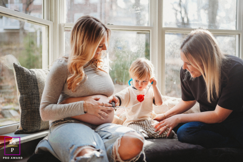 Bathed in Boston window light, this maternity scene quietly blends anticipation with childhood curiosity as a young child examines the baby bump like a tiny doctor at work. The intimate framing and soft tones make the connection feel beautifully natural.