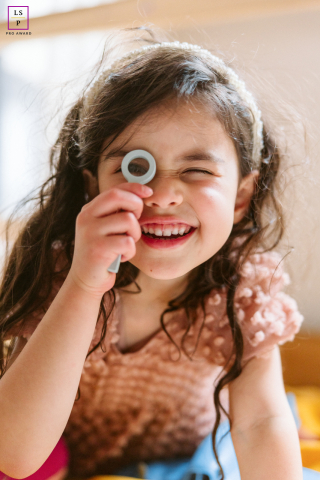 A playful portrait captures a young girl in Chicago, Illinois, peeking through a magnifying glass with a bright smile. Her curious expression and lively energy bring out a fun, imaginative personality filled with wonder and charm.