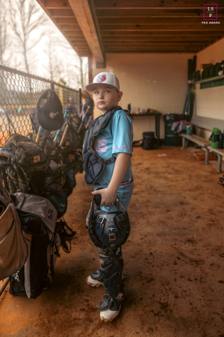 In Rome, Georgia, the young catcher stands quietly in the dugout holding his helmet as warm evening light spills across the dirt floor. The stillness and focused expression give the portrait determination, anticipation, and youthful confidence.