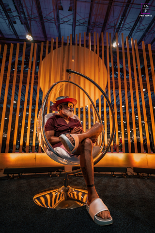 In New Orleans, Louisiana, the stylish man relaxes confidently in a hanging chair beneath glowing architectural lines and warm light. The bold composition and modern setting give the portrait personality, and a striking sense of contemporary style.