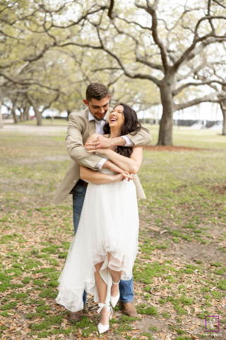 In Charleston, South Carolina, he wraps his arms around her from behind as she bursts into laughter beneath the sprawling trees. The relaxed movement and soft natural setting give the portrait warmth, romance, and playful intimacy.