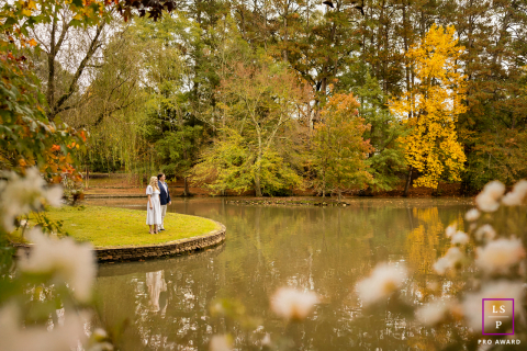 In Atlanta, Georgia, the couple stands quietly along the edge of the small island as autumn colors reflect across the still pond. The wide composition and soft foreground framing create a portrait filled with calm, romance, and seasonal beauty.