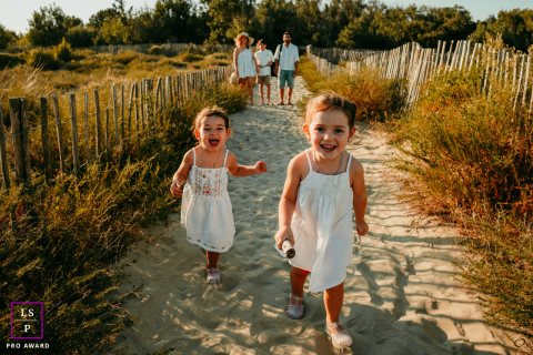 In La Grande Motte, France, the two young girls race excitedly toward the beach while their parents follow behind along the sandy path. The golden evening light and movement give the portrait warmth, freedom, and carefree summer energy.