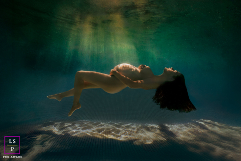Beneath the water, the expectant mother floats peacefully as soft light ripples across her body and the pool floor below. The minimal composition and weightless pose create a maternity portrait filled with calm and quiet beauty.