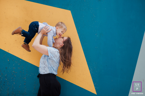 In Metz, the mother lifts her son high into the air as both share a moment of complete joy and trust. The bold geometric background adds graphic energy to the portrait, turning a playful interaction into something vibrant and wonderfully modern.