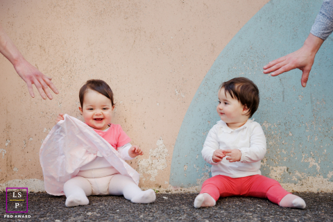 The Paris twin girls sit just out of reach while their parents’ hands enter the frame in a playful attempt to catch them. The simple backdrop and mirrored poses give the portrait humor, balance, and the sweet unpredictability of early childhood.