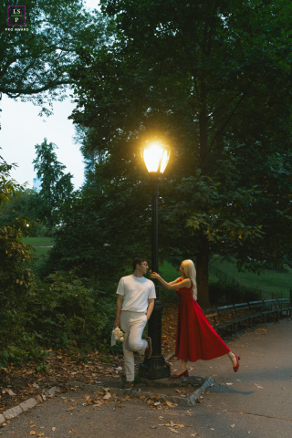 In Central Park, New York City, the couple shares a playful moment beneath the glow of a lone streetlamp as her red dress lifts with movement and energy. The surrounding darkness and towering trees give the portrait romance, motion, and cinematic charm.