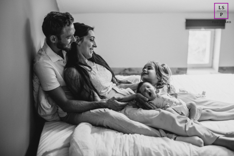 A family relaxes together on a bed as their young daughter laughs beside her sleepy baby sibling in Loire, France.