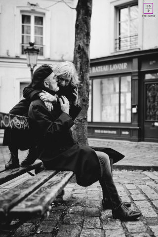 Older Couple Shares Affectionate Embrace On Cobblestone Street In Paris An older couple shares an affectionate embrace while sitting together on a bench along a quiet cobblestone street in Paris, France.
