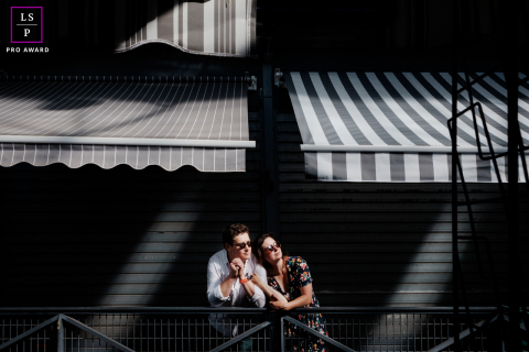 A couple leans closely together beneath striped awnings, surrounded by dramatic patterns of sunlight and shadow along a Parisian street.