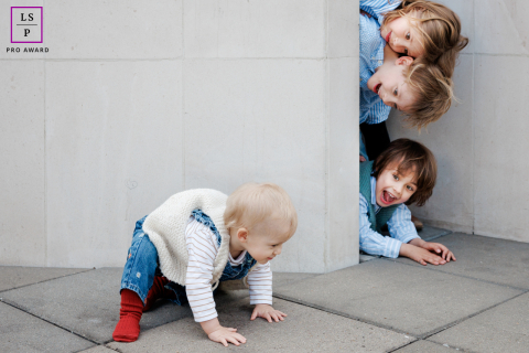 A young child crawls forward while three older siblings peek playfully around a wall during a lively family session in Paris.