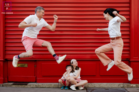 Parents leap playfully above their two seated children, creating a lively family scene against a bright red storefront in Paris.