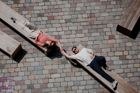 A couple relaxes on separate wooden benches, reaching toward each other beneath bright sunlight in a graphic overhead scene in Paris.