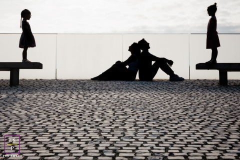 A family forms playful silhouettes against the bright sky, with parents seated between their daughters on benches overlooking Biarritz.