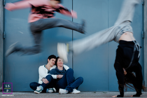 Two women sit together watching their teenagers leap and tumble playfully through the frame during a lively family session in Paris.