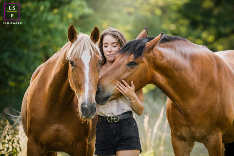 In Lyon, France, the young woman stands quietly between her two horses as they lean gently toward her. The soft evening light and close composition create a portrait filled with tenderness, connection, and the calm rhythm shared between animals and owner.