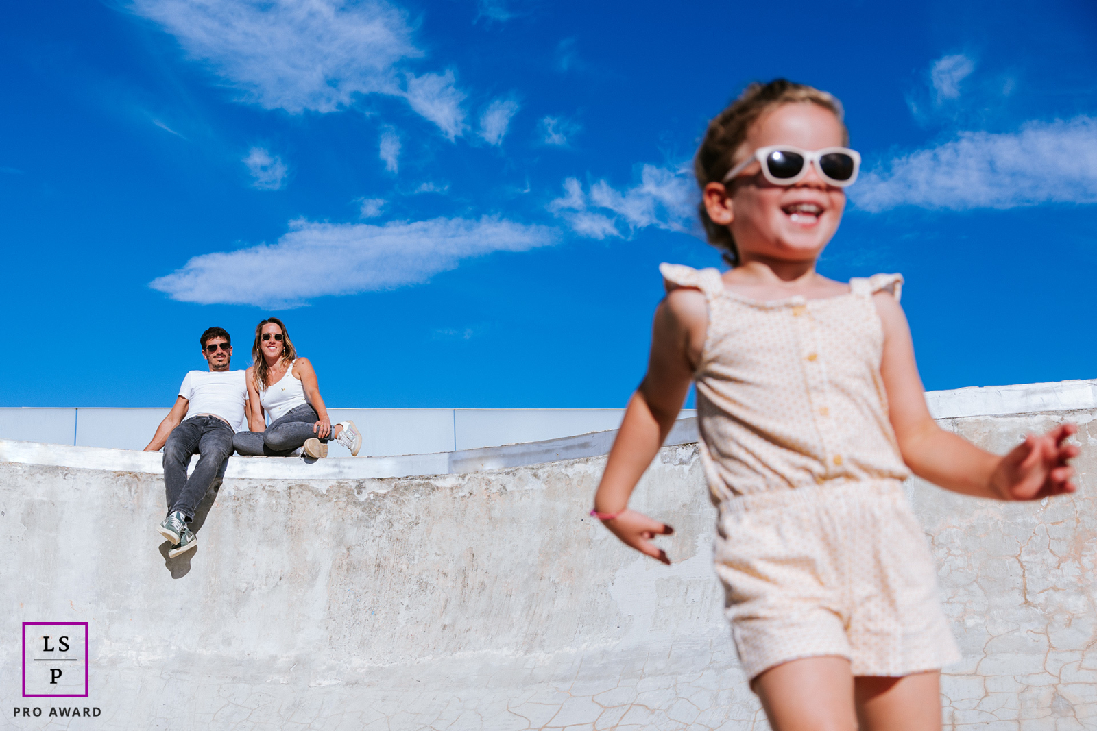 In Biarritz, this child races through the foreground in sunglasses while her parents relax quietly behind her beneath the vivid blue sky. The exaggerated perspective and playful movement give the photograph humor, freedom, and lively summer energy.