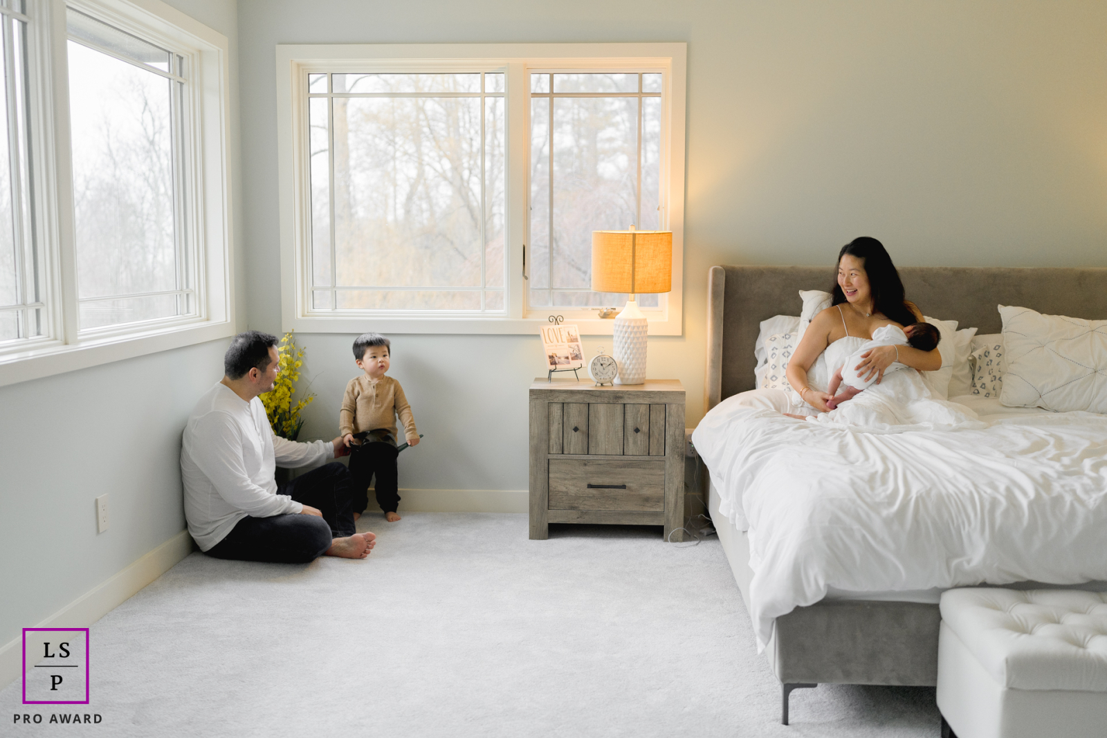 In NYC, the quiet rhythm of a growing family unfolds across the softly lit room as a mother cradles her newborn while father and son share a gentle moment nearby. The wide composition and calm tones give the portrait warmth, balance, and intimacy.
