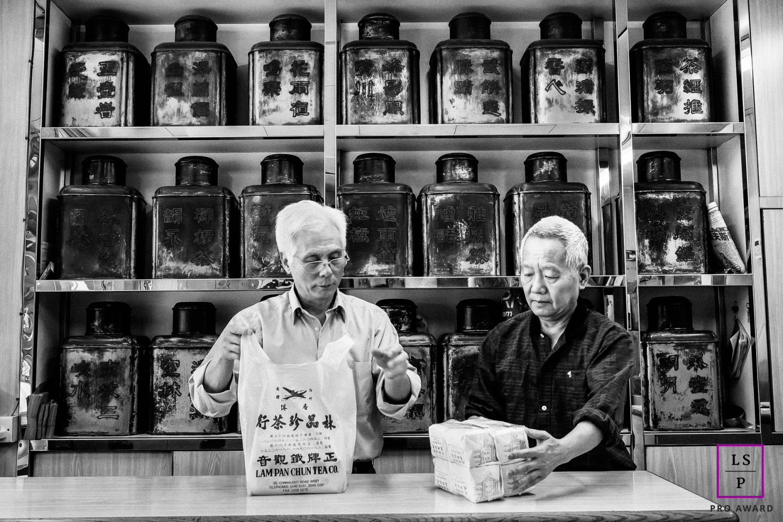 Portrait professionnel de deux hommes de la Lam Pan Chun Tea Company à Hong Kong, capturé dans un cliché saisissant en noir et blanc alors qu'ils emballent avec diligence les commandes. Cette image met en valeur la nature travailleuse et dévouée de l'entreprise.