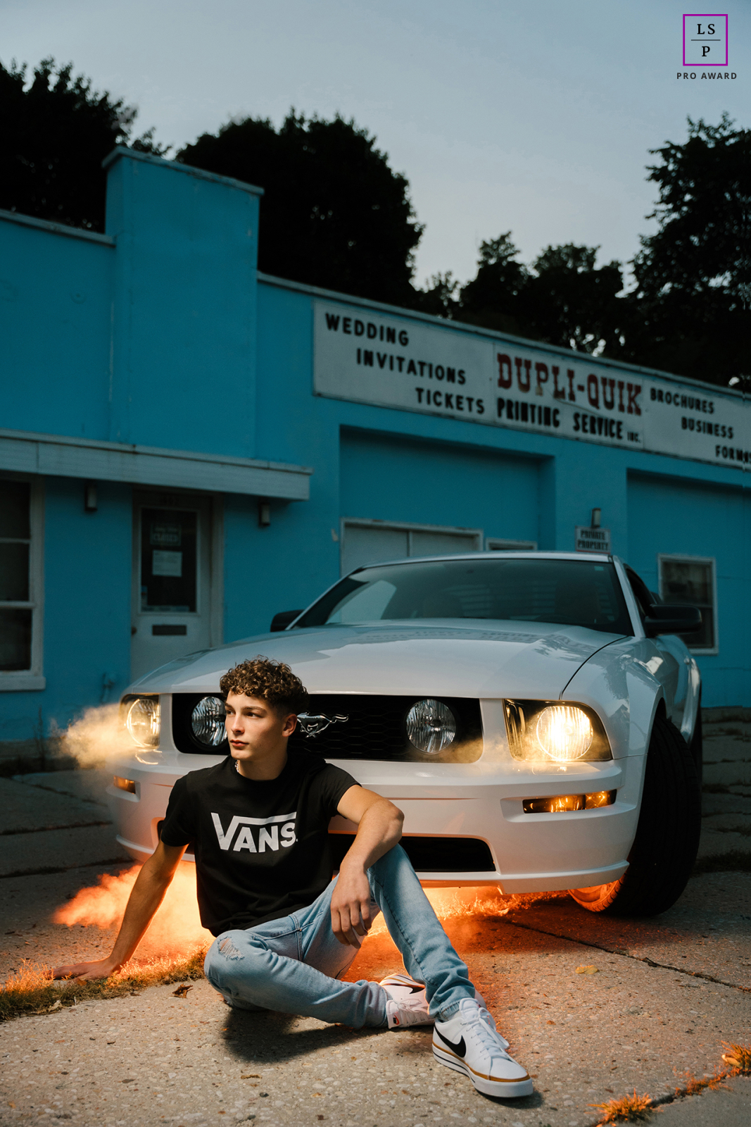 A senior posing with his new Ford Mustang in West Bend, WI. He sits on concrete with headlights on at dusk, as smoke rises from under the car.