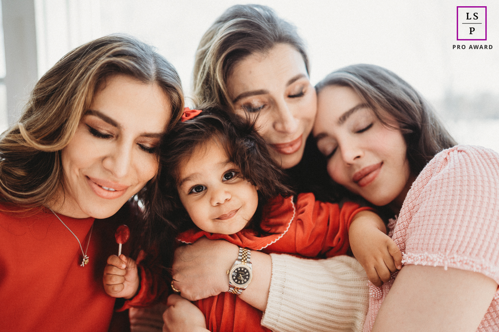 In Boston, Massachusetts, a young toddler girl holding a red lollipop looks at the camera, embraced by three women with their eyes closed, symbolizing generations of love.