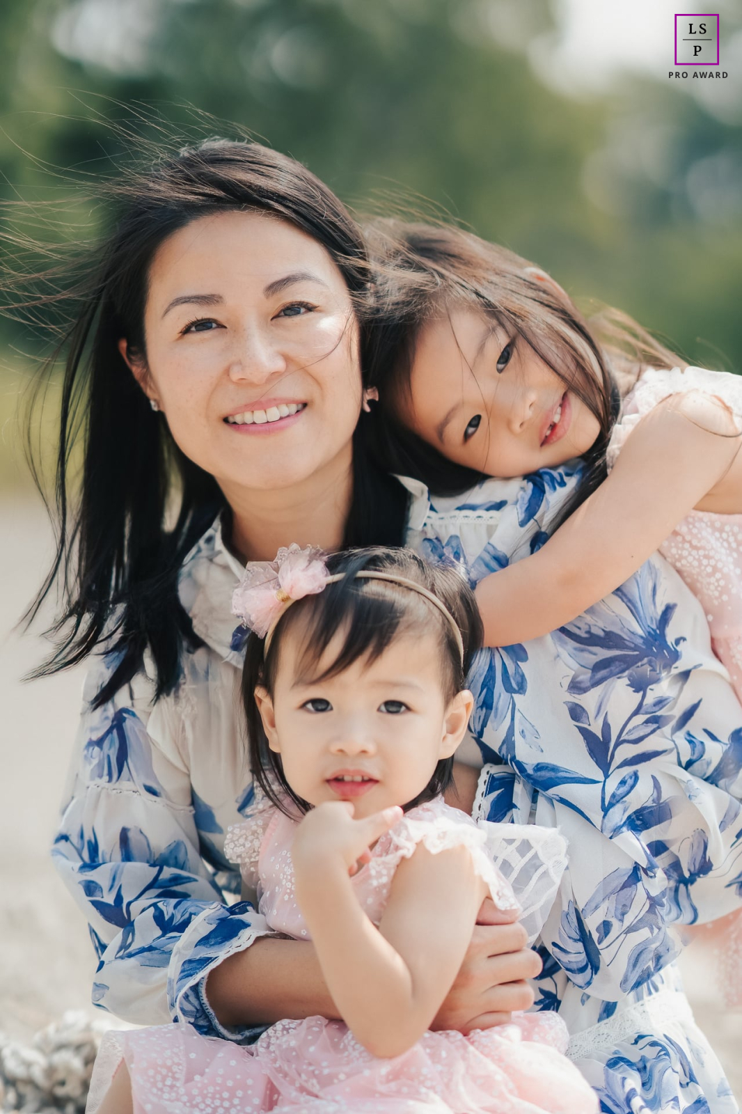 In Toronto, this mother gathers both daughters into one affectionate embrace as the older child leans gently across her shoulder. The soft outdoor light and close arrangement give the photograph warmth, tenderness, and beautifully natural connection.