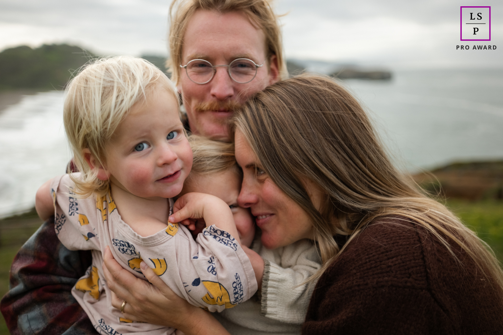 At Rodeo Beach in Marin, this family squeezes together against the ocean backdrop while dad and son meet the camera directly. The tight grouping and soft coastal light give the photograph warmth, intimacy, and playful family closeness.