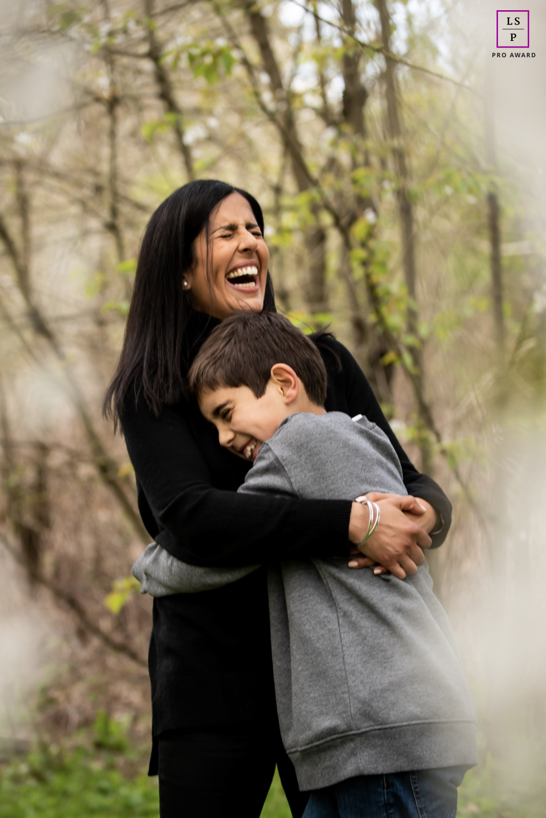 Among the soft spring trees in St Albans, a mother bursts into laughter while holding her son tightly against her. The blurred foreground and natural expressions give the photograph warmth, movement, and the unmistakable comfort of family closeness.