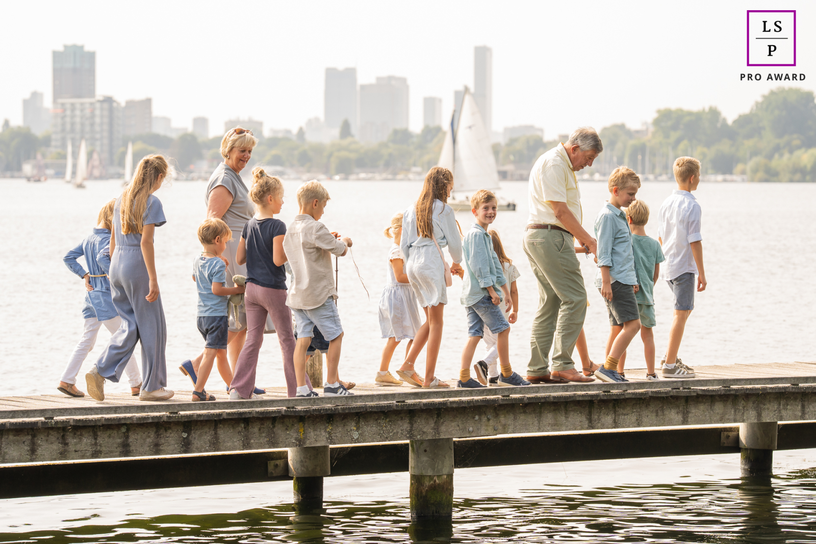 Along the dock at Kralingse Plas in Rotterdam, grandparents lead a parade of grandchildren beside the water, each child adding their own rhythm to the scene. The layered line of figures and distant skyline give the image wonderful energy and connection.