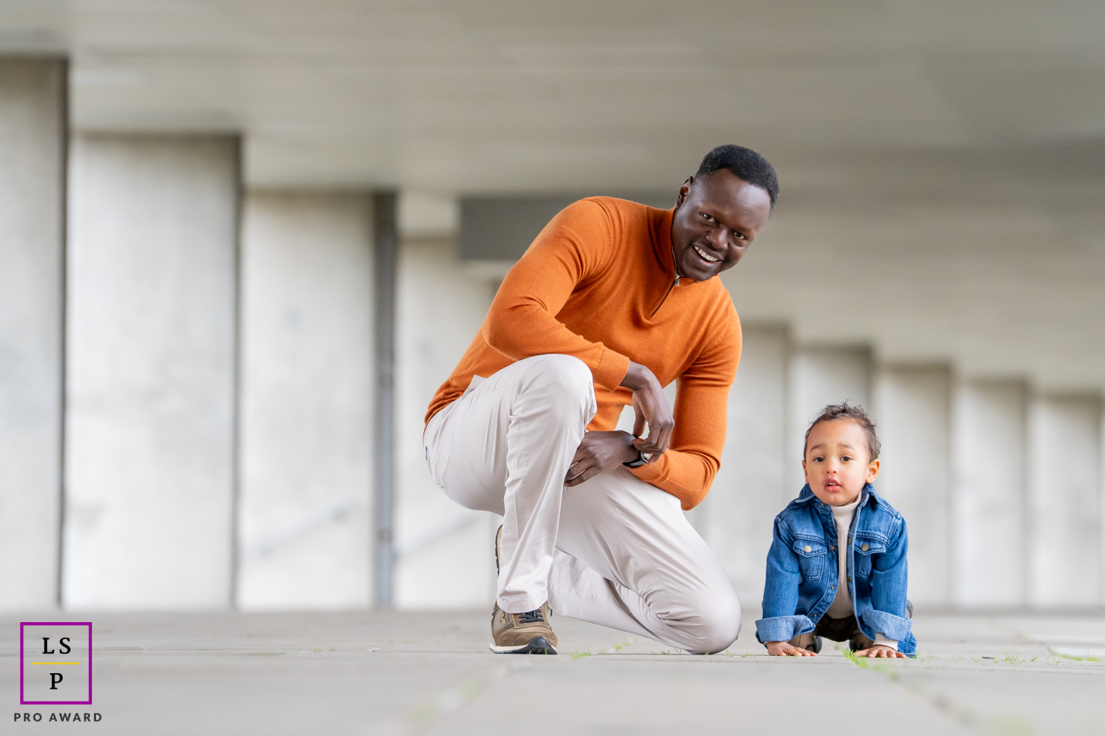 At the Dutch Architect Institute in Rotterdam, a father crouches beside his young son as the child studies the ground with total concentration. The repeating concrete lines and open space give this tender family photograph a striking modern simplicity.