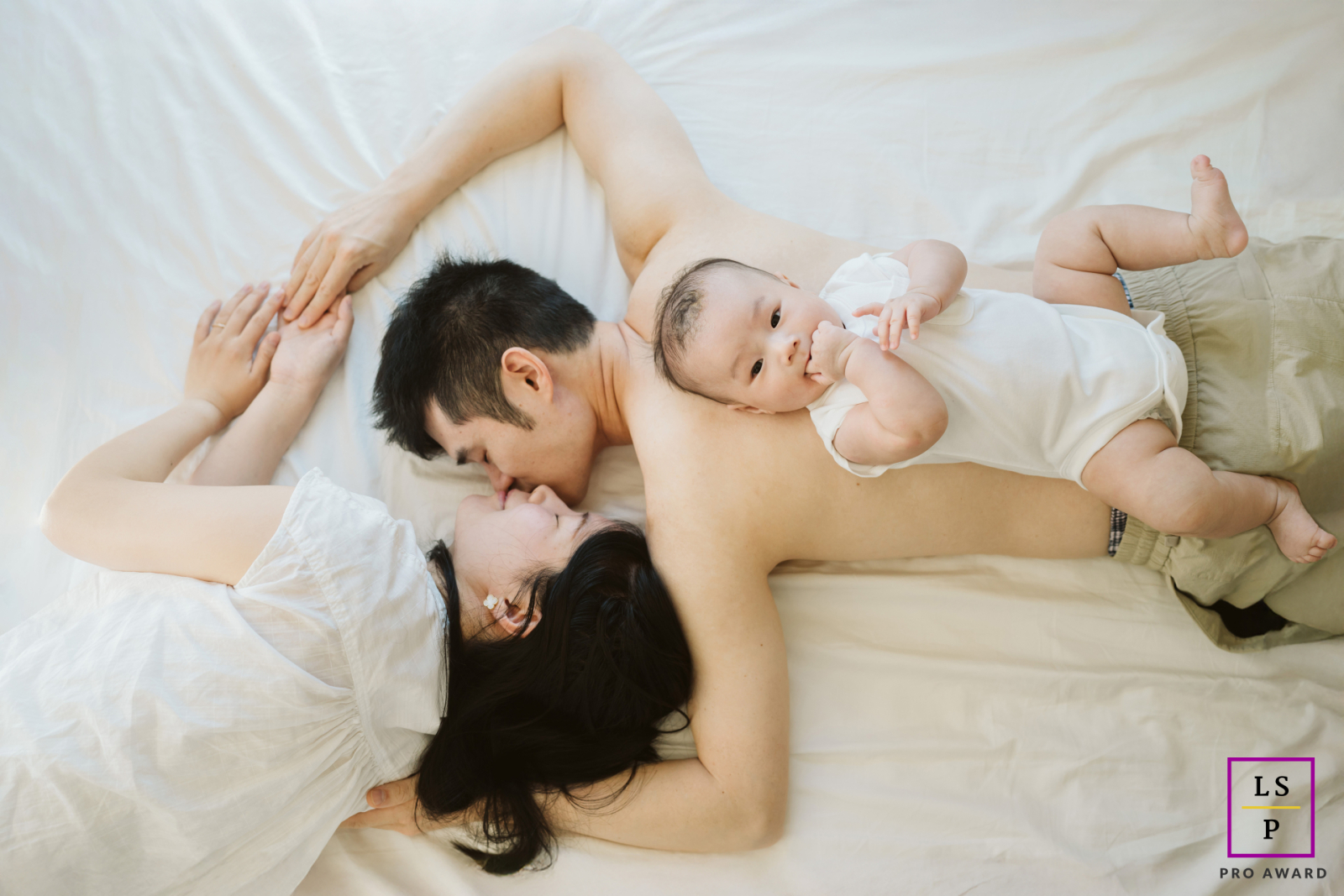 In Jersey City, a baby sprawls comfortably across dad’s back while the parents share a quiet kiss beneath him. The overhead composition and soft white tones turn this playful family arrangement into something intimate, tender, and unexpectedly poetic.