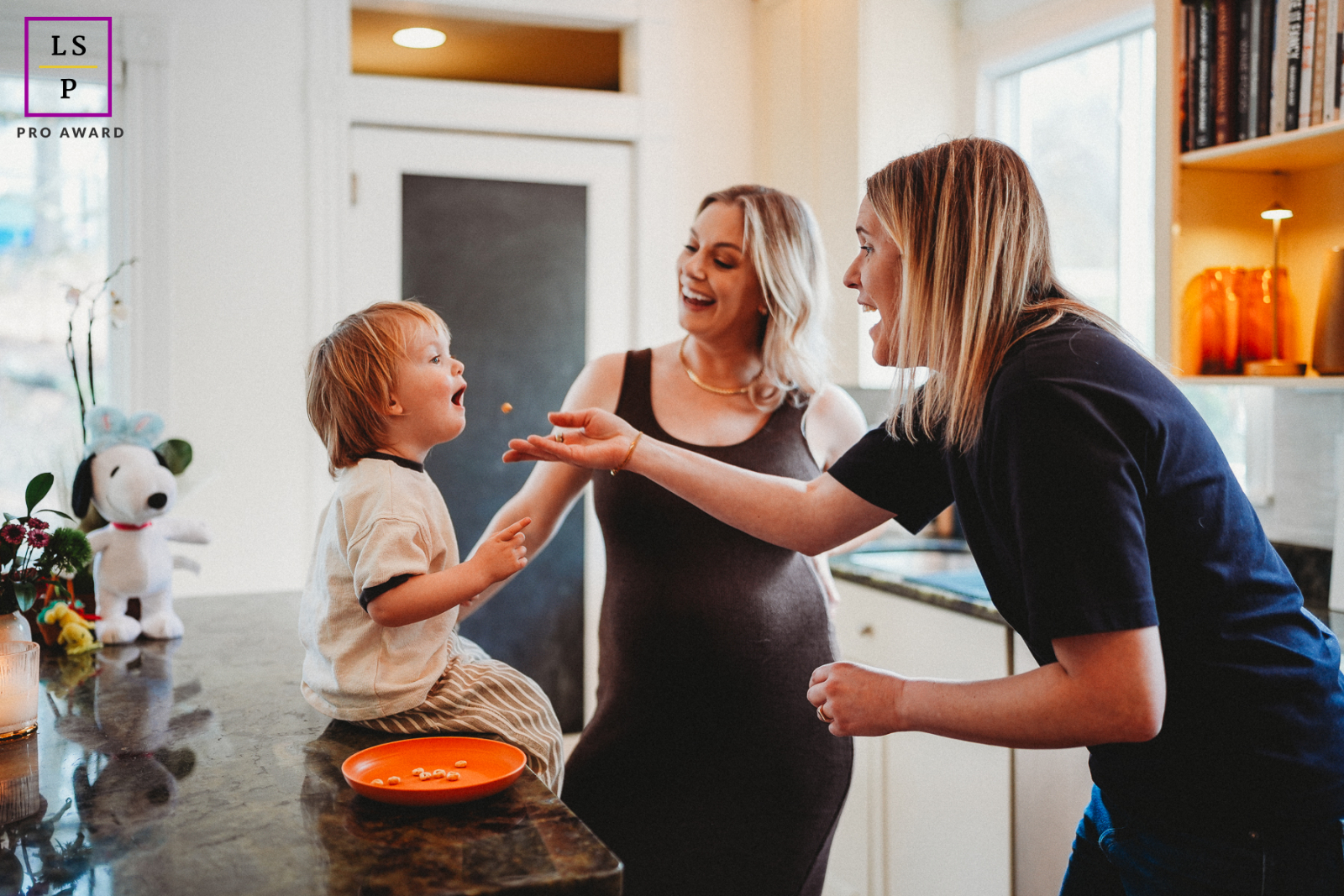 In this lively Boston kitchen, a toddler proudly offers Cheerios while two adults erupt with encouragement and laughter. The open gestures, bright interior light, and playful energy turn ordinary breakfast chaos into something wonderfully heartfelt.