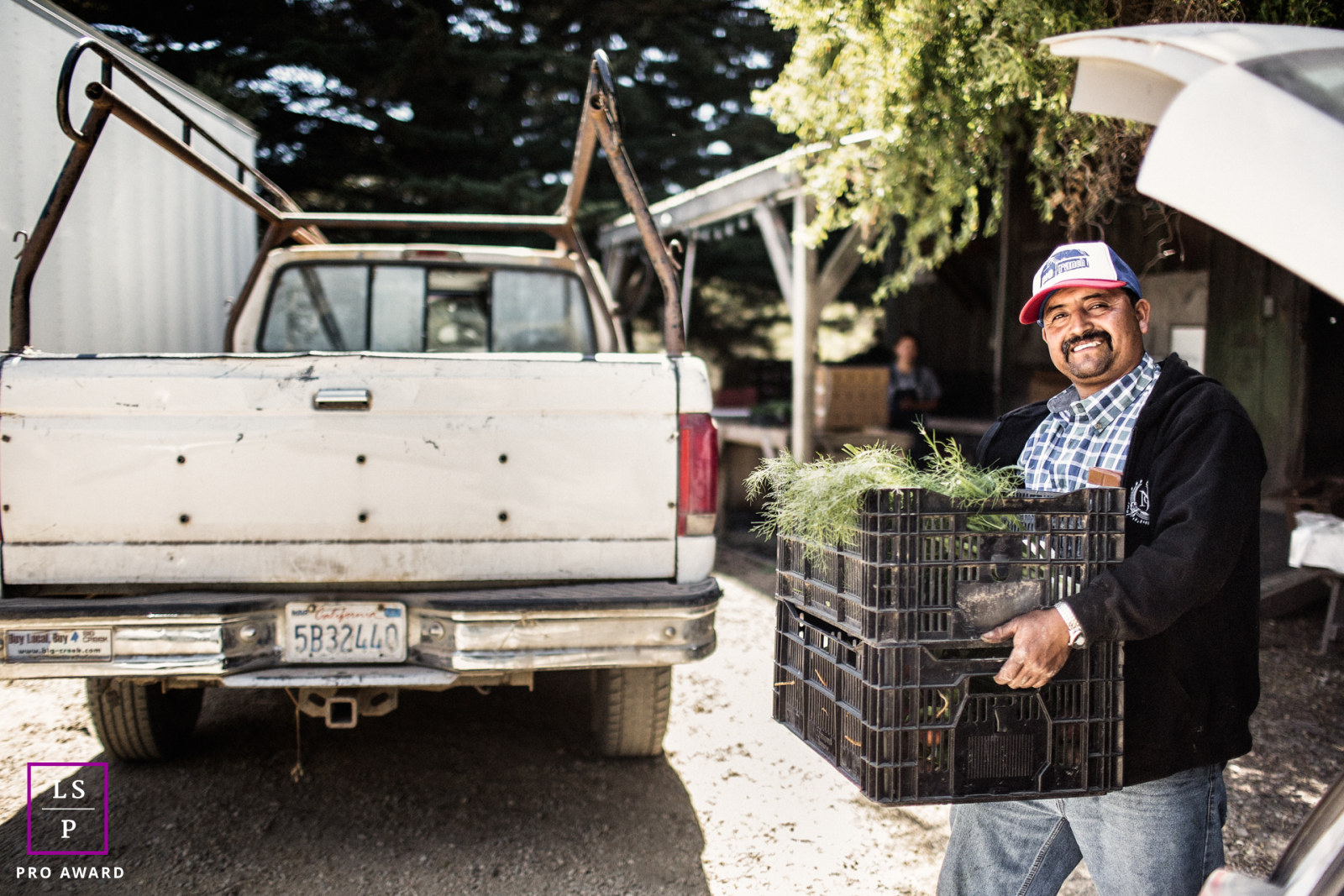 Portrait d'entreprise professionnel au Pie Ranch, Pescadero, Californie : un homme souriant transportant des légumes frais dans des caisses en plastique jusqu'à un camion de travail, représentant l'éthique de la ferme à la table de cet établissement côtier.