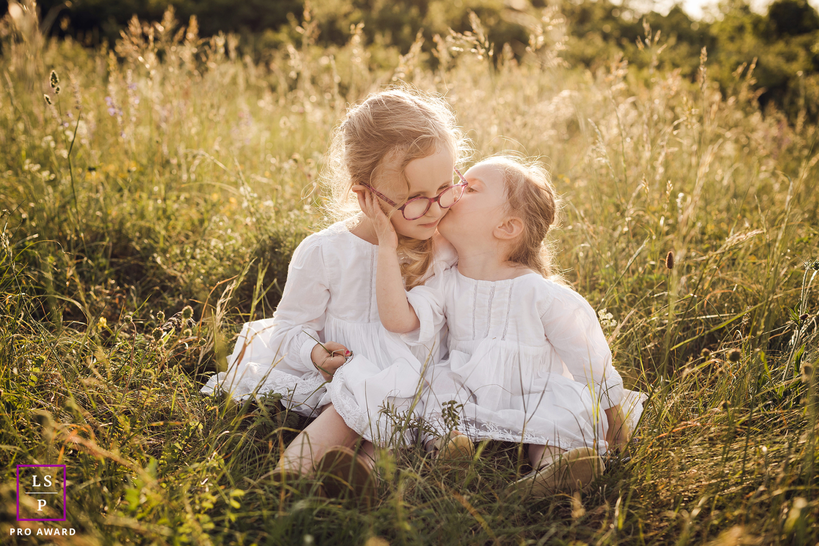 In Lyon, France, two sisters sit in the grass, with the younger sister sweetly kissing her older sister on the cheek, bathed in sunlight amidst the beauty of nature.