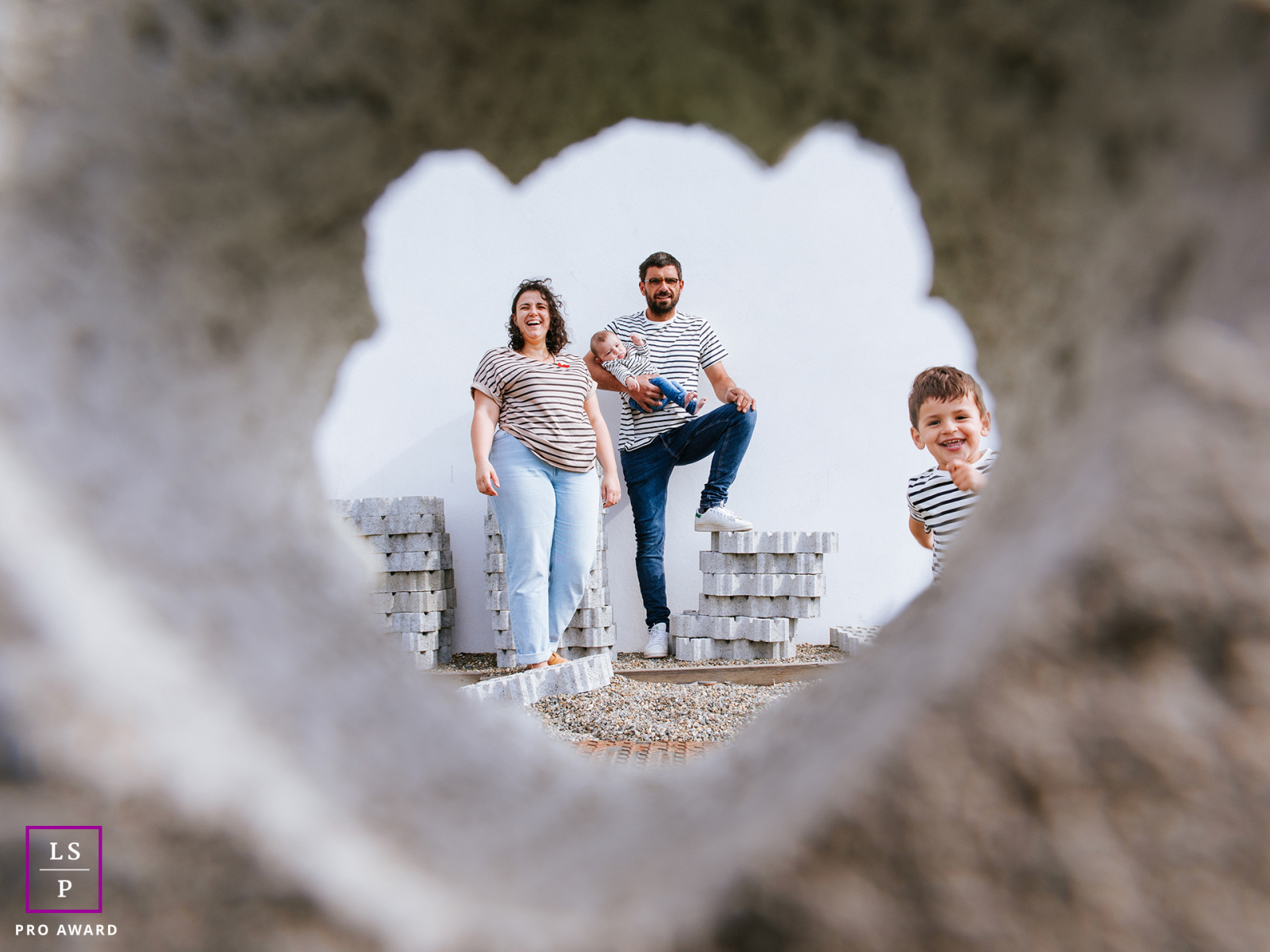 Framed through a heart shaped opening in Bardos, this family appears surrounded by humor, love, and unfinished construction. The playful perspective and unexpected peeking child give the photograph charm, creativity, and lighthearted storytelling.