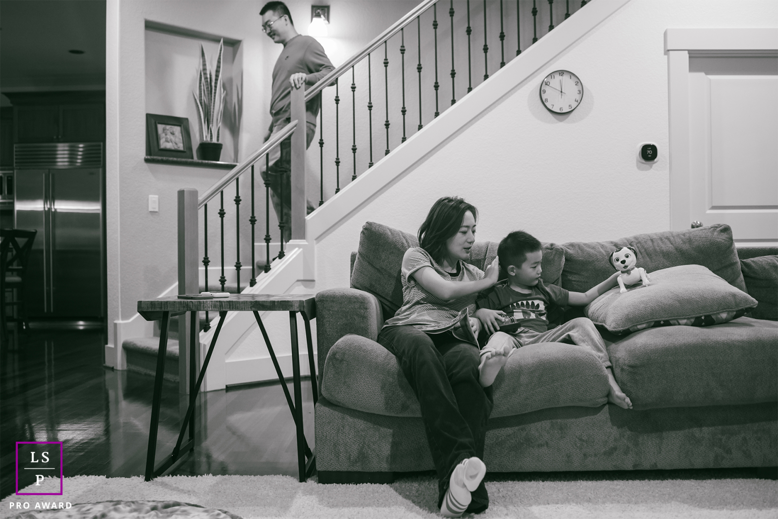 In this Oregon home, everyday family life unfolds quietly as a mother reads with her son while the father descends the stairs after bedtime duties. The black and white tones give the candid scene warmth, stillness, and honesty.