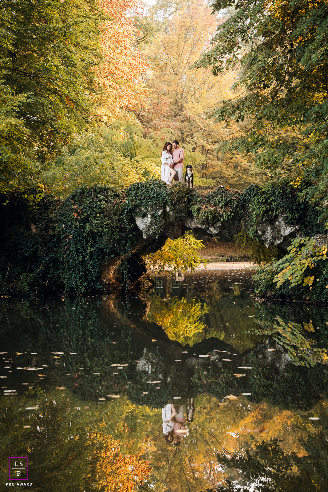 Surrounded by autumn color in Lyon, this expecting couple stands with their dog atop a vine covered bridge reflected perfectly below. The layered symmetry and peaceful landscape give the photograph romance, stillness, and storybook charm.