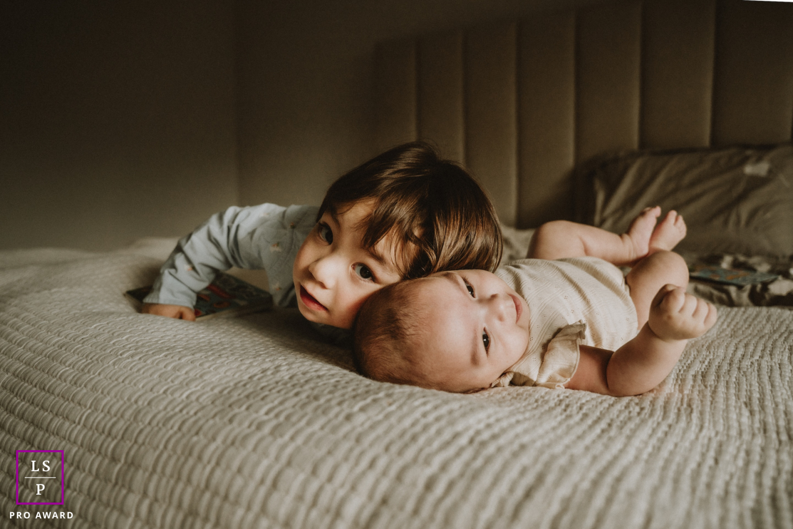In this softly lit Toronto bedroom, a young brother presses close beside his baby sister with effortless tenderness. The warm shadows and low perspective make the photograph feel intimate, peaceful, and beautifully centered on quiet sibling connection.