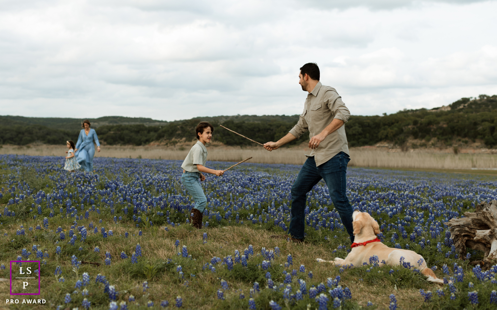 In Marble Falls, TX, a father and son duel with sticks among the bluebonnets while mother and daughter wander peacefully in the distance. The wide landscape and layered family moments give the portrait humor and the relaxed rhythm of togetherness.
