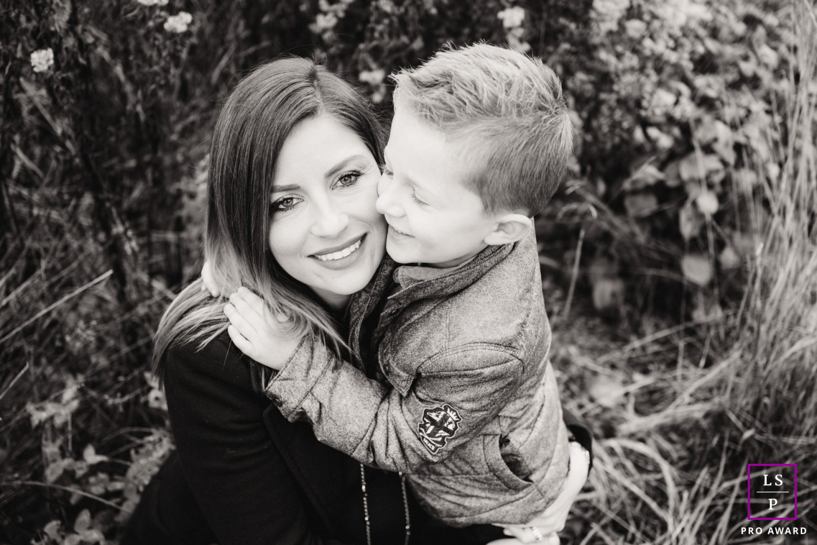 In Alsace, a black-and-white shot captures a toddler boy kissing his squatting mom on the cheek, highlighting their loving bond.