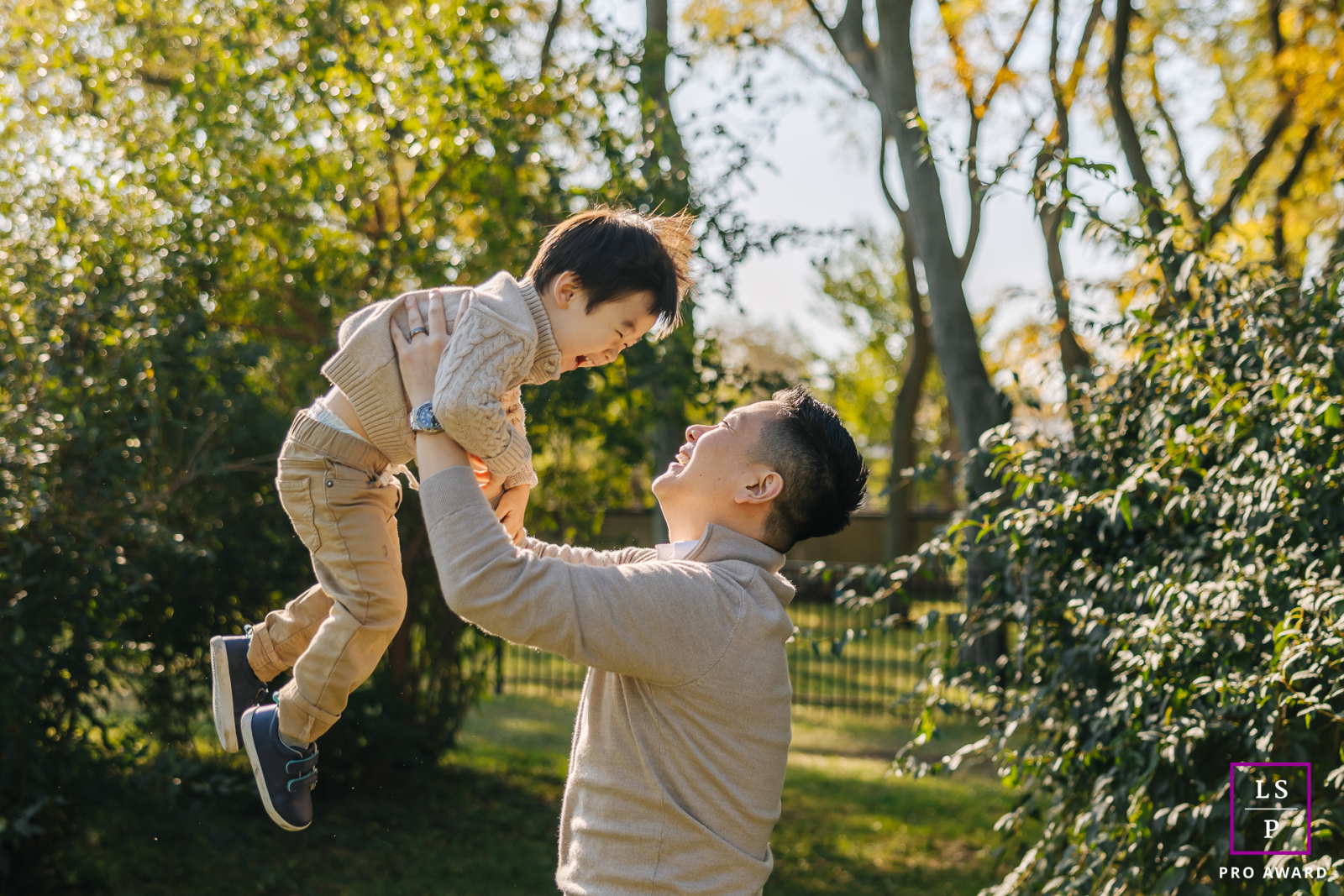 In Chicago, IL, under golden sunlight, a dad joyfully lifts his son outdoors, surrounded by nature and laughter.